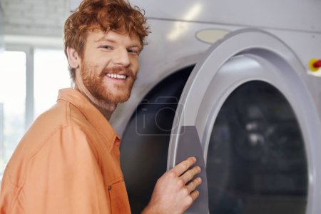 sonriente joven pelirrojo hombre mirando a la cámara cerca de la lavadora en auto servicio de lavandería