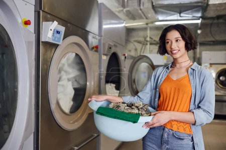 Lächelnde junge Asiatin hält Korb mit Kleidung neben Waschmaschine in Münzwäsche