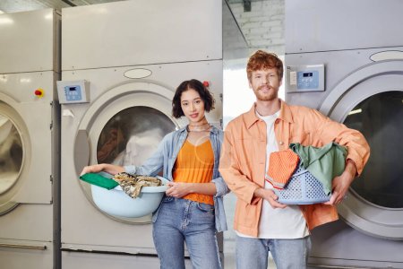 smiling interracial couple holding basins near washing machines in coin laundry