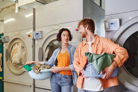 positive interracial couple holding clothes near washing machines in coin laundry