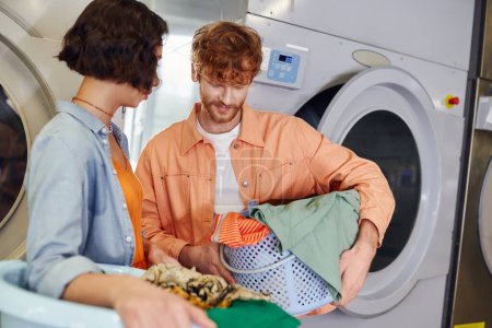 junger rothaariger Mann hält Waschbecken mit Kleidung in der Nähe asiatischer Freundin in Münzwäsche