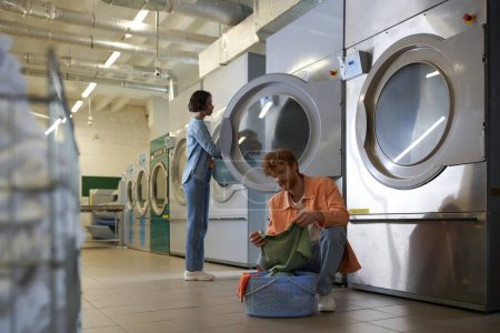 young man holding clothes near basket and asian girlfriend in public coin laundry
