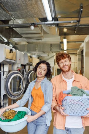 pareja interracial positiva sosteniendo cestas con ropa y mirando a la cámara en la lavandería de monedas