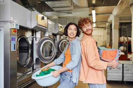 smiling multiethnic couple standing back to back and holding basins with clothes in coin laundry
