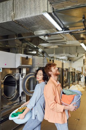 joyful interracial couple holding basket with clothes and standing back to back in public laundry