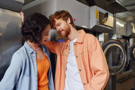 young redhead man flirting with asian girlfriend near washing machines in coin laundry