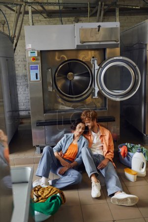 romantic interracial couple sitting on floor near clothes and washing machine in public laundry