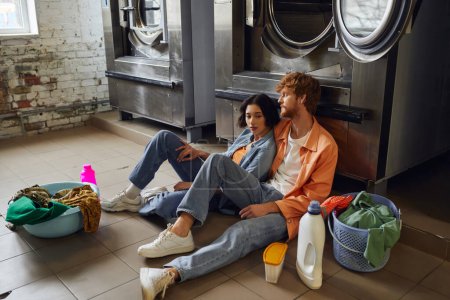 romantic multiethnic couple sitting near clothes and detergents on floor in public laundry