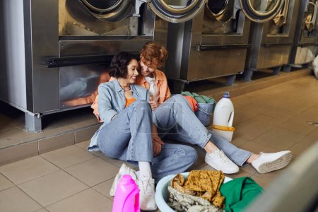 smiling man hugging asian girlfriend near clothes and detergent on floor in public laundry