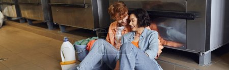 pareja multiétnica positiva sentada cerca de la ropa y el detergente en el suelo en la lavandería moneda, pancarta