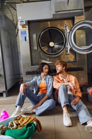 young interracial couple sitting near clothes and detergent on floor in public laundry
