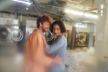 smiling young man hugging asian girlfriend while standing in blurred coin laundry