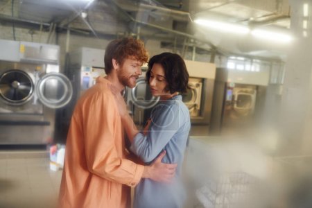 cheerful redhead man hugging young asian girlfriend in blurred public laundry
