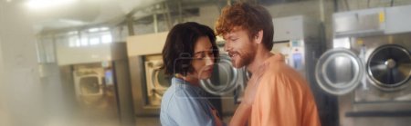 smiling young redhead man standing near brunette asian girlfriend in coin laundry, banner