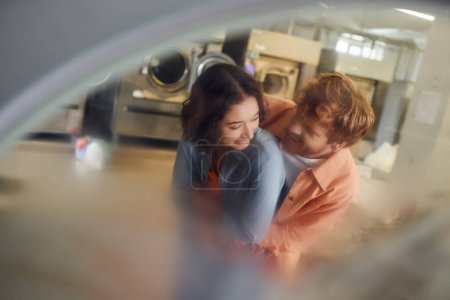 young multiethnic couple hugging while standing near blurred washing machines in coin laundry