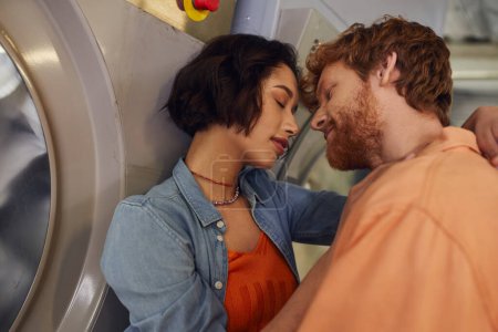 young multiethnic romantic couple kissing near washing machine in public laundry