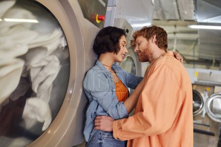 smiling asian woman kissing redhead boyfriend near washing machine in public laundry