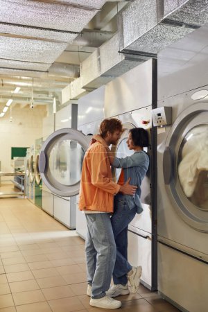widok z boku młody romantyczny wieloetniczny para przytulanie w pobliżu pralek w praniu publicznym