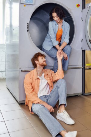 smiling asian woman holding hand of boyfriend while sitting on washing machine in coin laundry