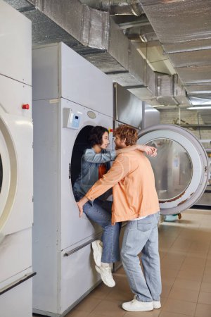 smiling young asian woman embracing boyfriend while sitting on washing machine in public laundry