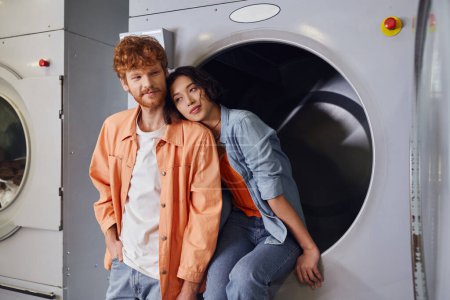 young asian woman sitting on washing machine near redhead boyfriend in public laundry