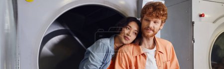 young asian woman standing near redhead boyfriend near washing machine in coin laundry, banner