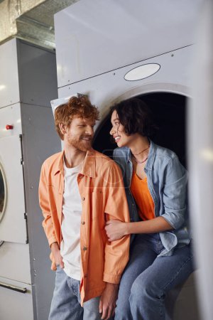 cheerful young asian woman hugging redhead boyfriend while sitting on washing machine in laundry