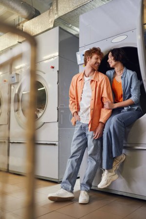 Téléchargez les photos : Pleine longueur de joyeux couple multiethnique parlant près de la machine à laver dans la buanderie publique - en image libre de droit