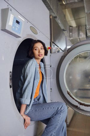 joven morena asiática mujer sentado en lavadora en público lavandería