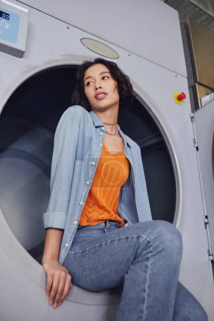 ángulo bajo vista de la joven mujer asiática mirando hacia otro lado mientras está sentado en la lavadora en la lavandería pública