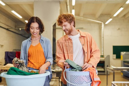 smiling young multiethnic couple putting clothes in baskets in public laundry on background