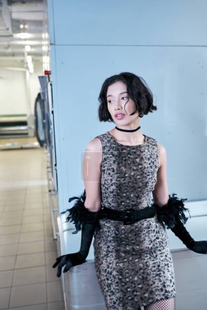 fashionable young asian woman in dress and gloves standing near wall in pubic laundry