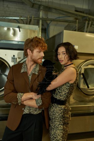 Photo for Fashionable young asian woman looking at camera near redhead boyfriend in public laundry - Royalty Free Image
