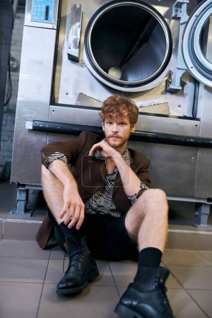 modischer Mann in Jacke und Shorts sitzt neben Waschmaschine in öffentlicher Wäscherei