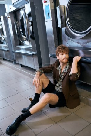 trendy redhead man in jacket posing near washing machine in public laundry