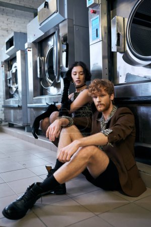 Selbstbewusster Mann in Jacke sitzt neben asiatischer Freundin in Kleid und Waschmaschine in öffentlicher Wäscherei