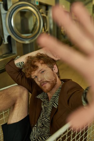 junger rothaariger Mann in Jacke und schickem Hemd blickt in die Kamera, während er im Wäschekorb in der Münzwäsche sitzt