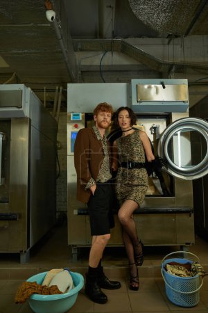 fashionable young interracial couple looking at camera near clothes in coin laundry
