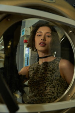 fashionable young asian woman standing behind glass of washing machine in coin laundry