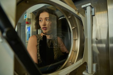 trendy young asian woman posing near door of washing machine in public laundry