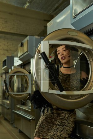 stylish young asian woman in dress posing near door of washing machine in coin laundry