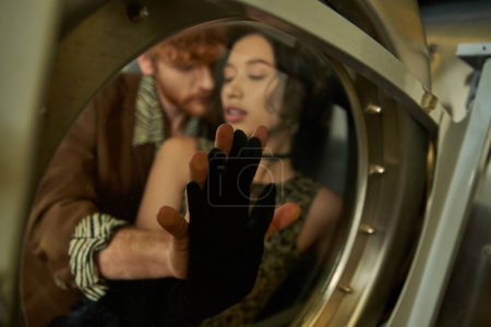 pareja multiétnica borrosa y elegante tocando el vidrio de la lavadora en la lavandería de monedas