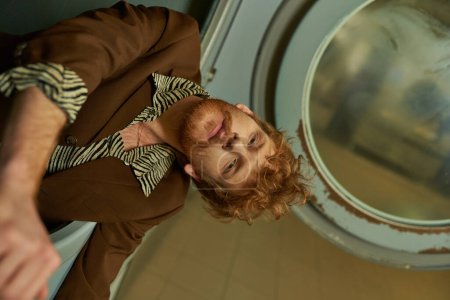 fashionable young redhead man in jacket posing in washing machine in public laundry