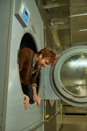 cheerful and trendy young redhead man posing in washing machine in public laundry