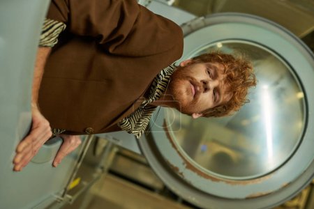 portrait of young fashionable redhead man in jacket posing in washing machine in coin laundry