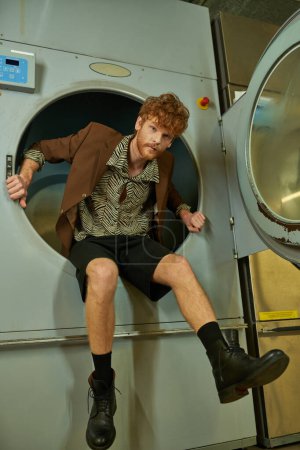 stylish redhead man in shorts and jacket looking at camera from washing machine in coin laundry
