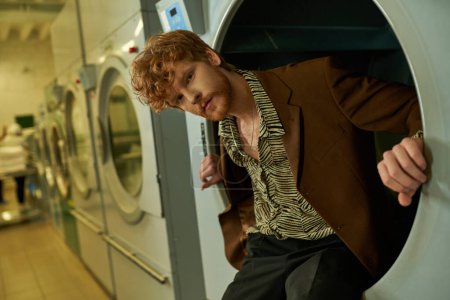 confident young redhead man looking at camera while posing in washing machine in coin laundry