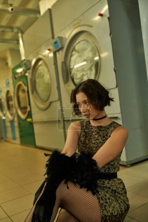 confident asian woman in dress and gloves with feathers posing in public laundry in evening