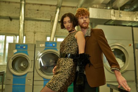fashionable asian woman in dress looking at camera near redhead boyfriend in self service laundry