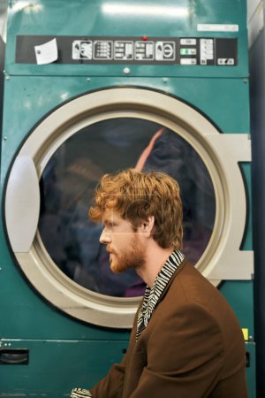 Foto de Vista lateral del joven pelirrojo de moda posando cerca de la lavadora en la lavandería pública - Imagen libre de derechos